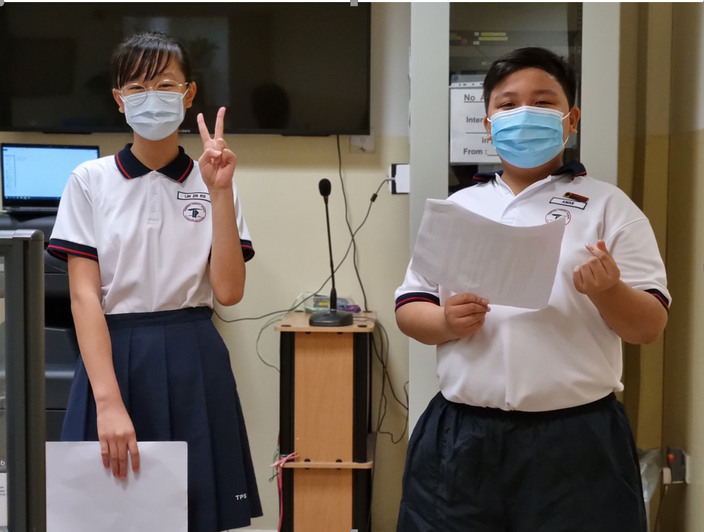 Two students in school uniforms and face masks stand holding papers, one making a peace sign.